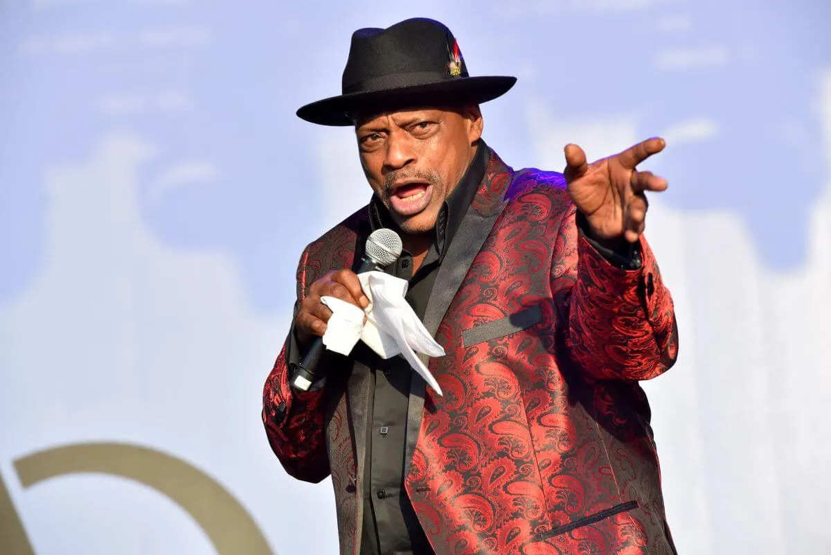 Alexander O'Neal wears a red, patterned suit and a fedora. He sings into a microphone.