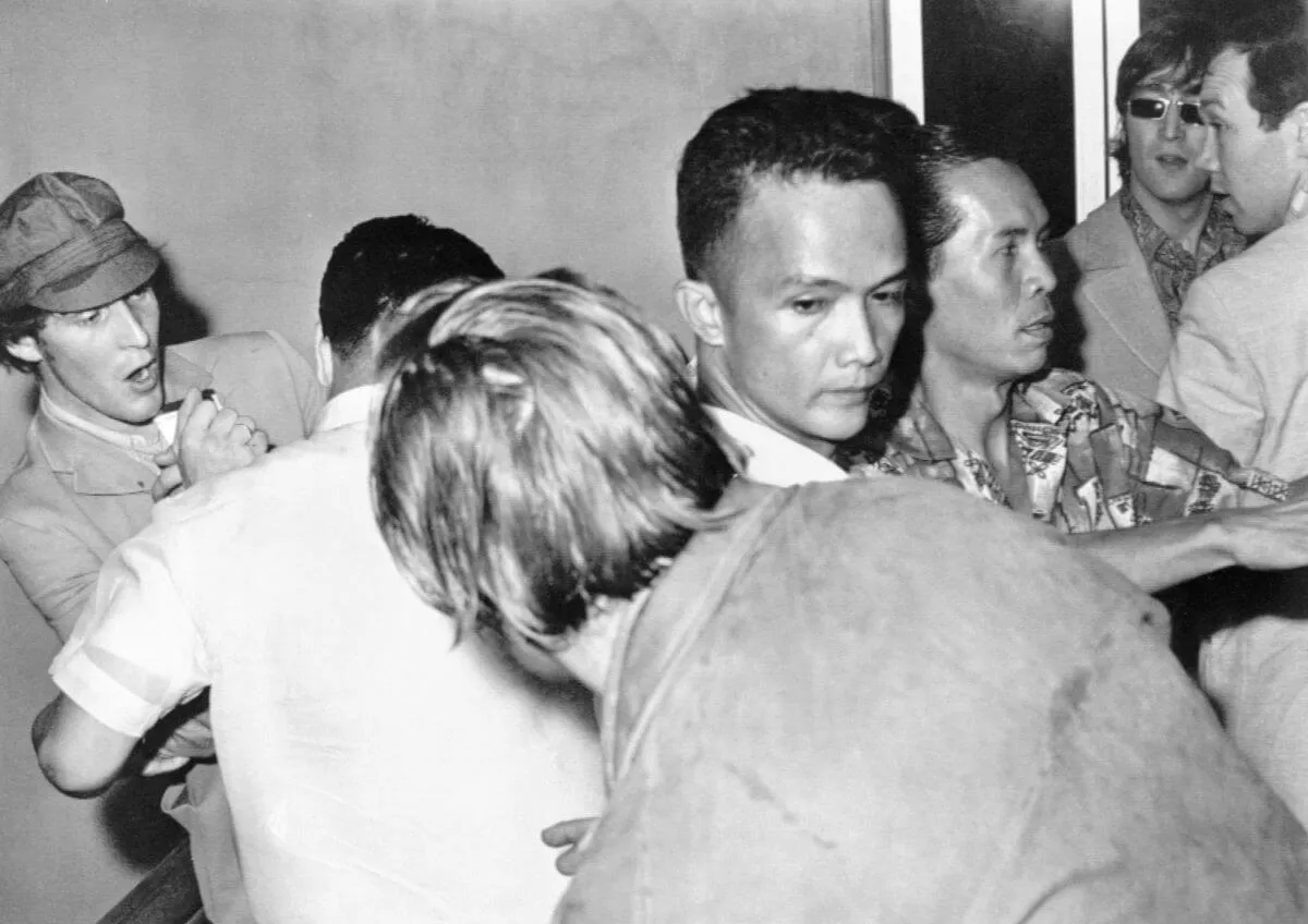 A black and white picture of John Lennon standing amid a crowd of people at the airport in the Philippines.