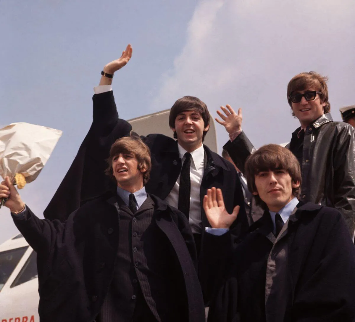 The Beatles wear suits and wave from the open door of an airplane.