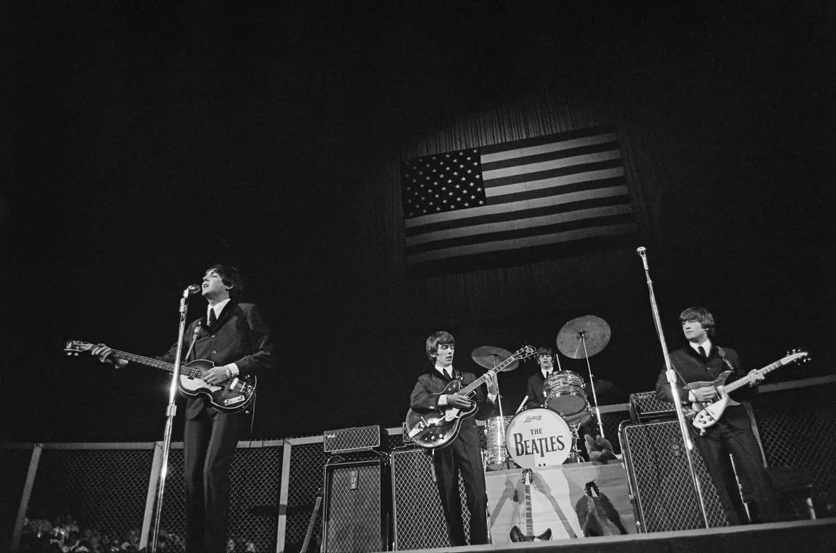 A black and white picture of The Beatles playing a concert on a stage. There is an American flag behind them.