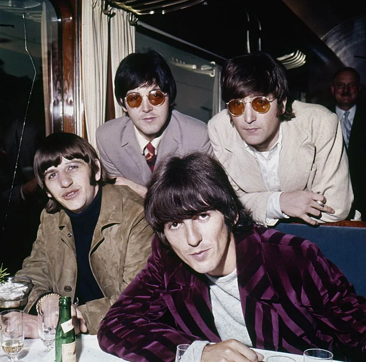 George Harrison, Ringo Starr, Paul McCartney, and John Lennon lean over a table. McCartney and Lennon wear circular, red-tinted glasses.