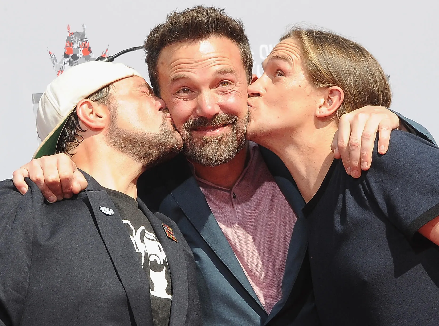 Kevin Smith and Jason Mewes kissing either side of Ben Affleck's cheeks as he holds on to their shoulders and smiles