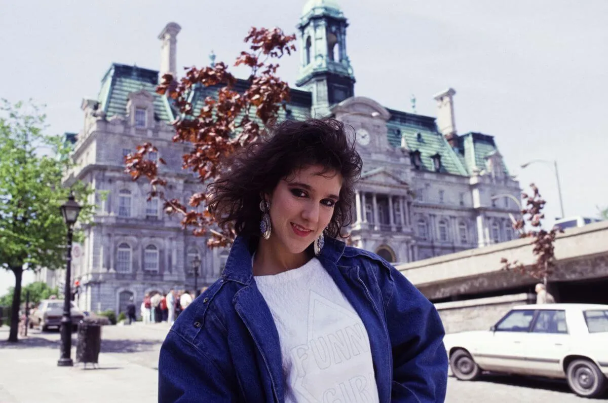 Celine Dion wears a denim jacket and white shirt. She poses outside in front of a building in Montreal.