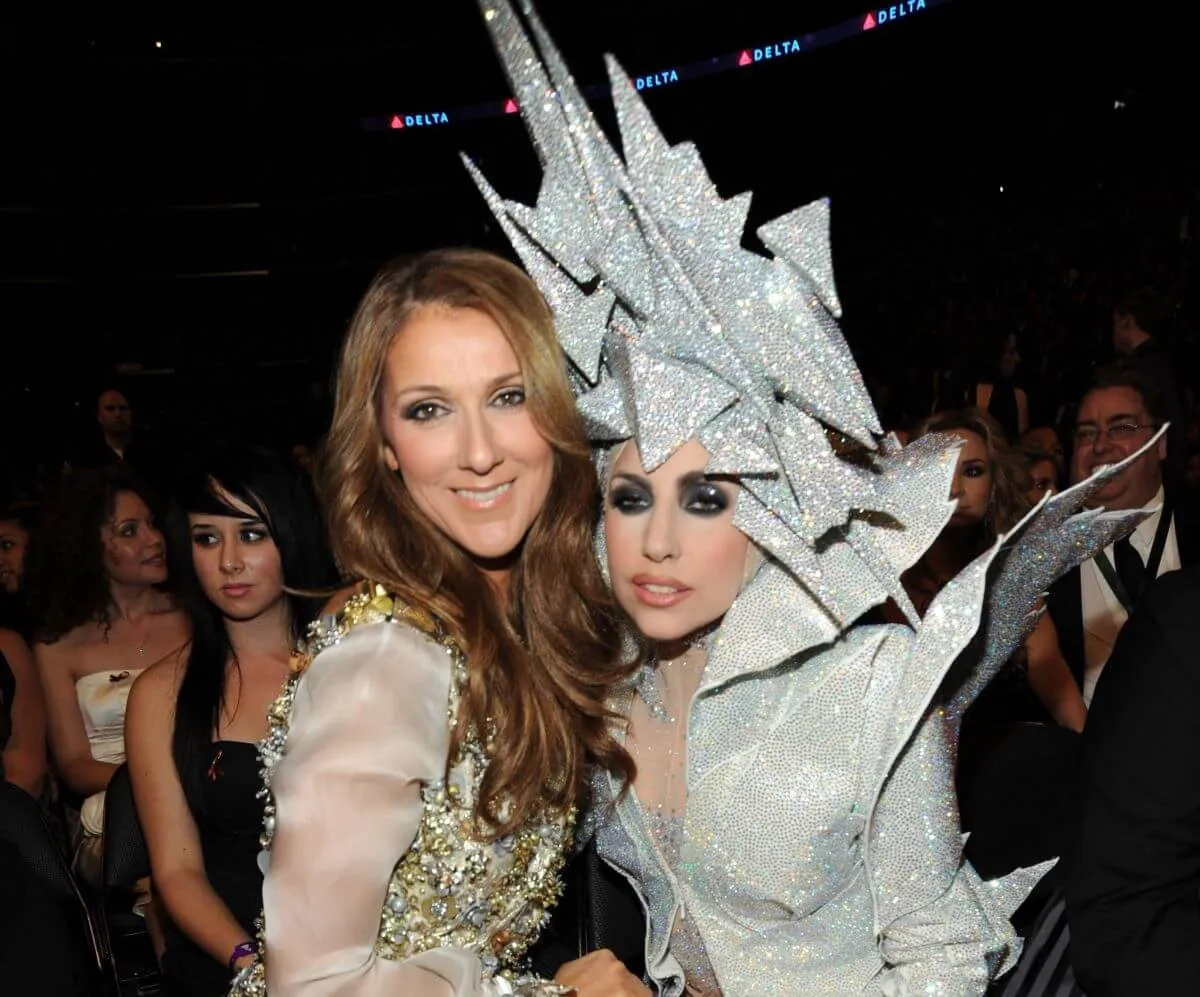 Celine Dion and Lady Gaga pose together. Dion wears a gold dress and Gaga wears a silver top and matching headpiece.