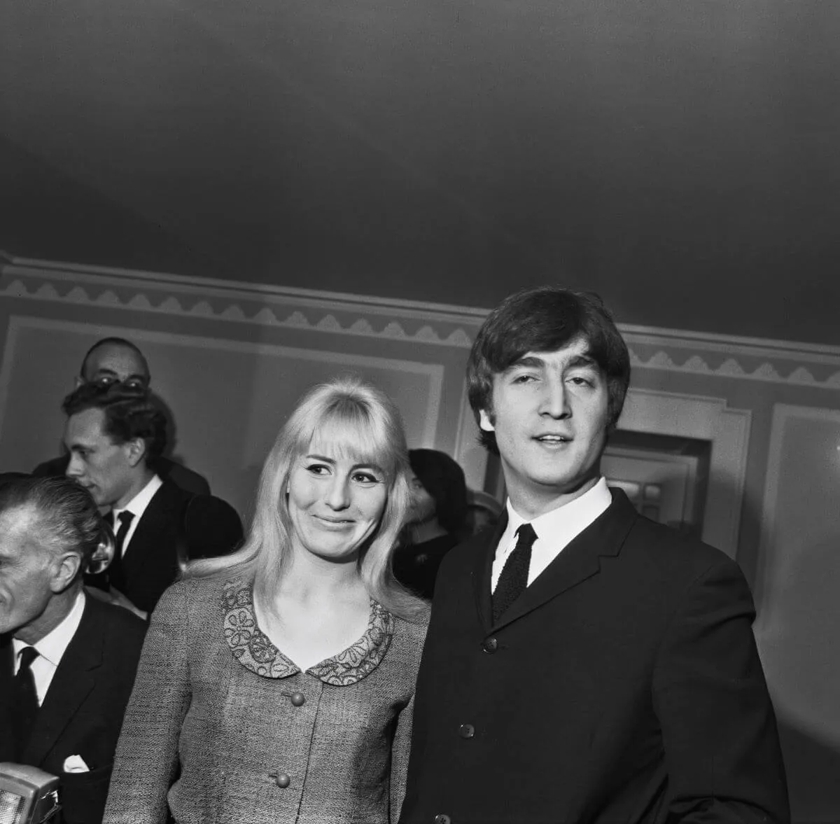 A black and white picture of Cynthia and John Lennon standing in a room together. There are a number of men behind them.