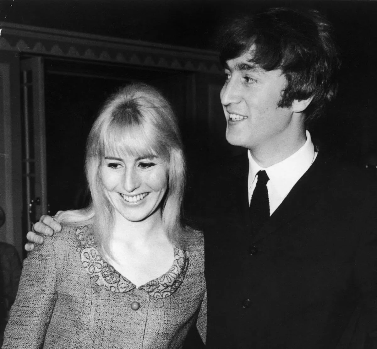 A black and white picture of John Lennon wearing a suit and standing with his arm around Cynthia Lennon's shoulders.
