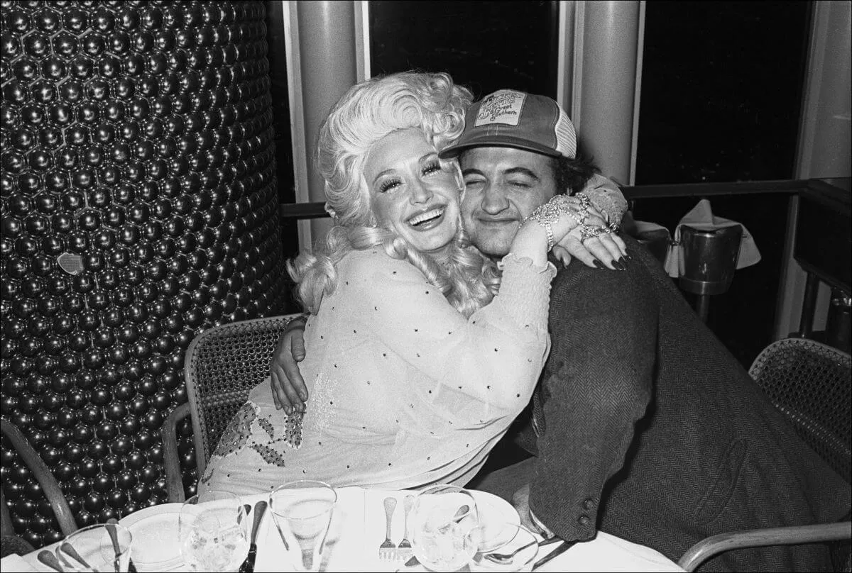 A black and white picture of Dolly Parton and John Belushi sitting at a table and embracing.