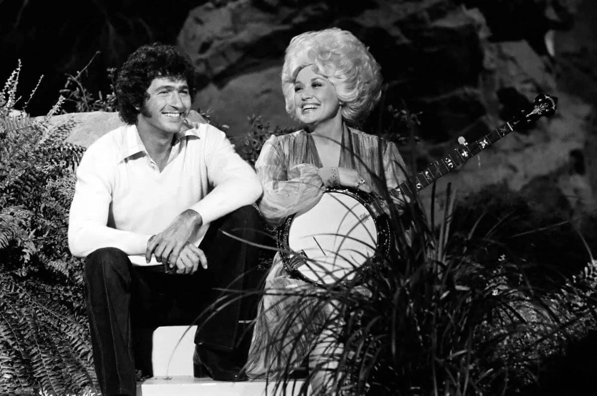 A black and white picture of Dolly Parton holding a banjo and sitting next to Mac Davis.