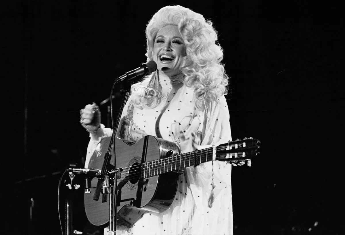 A black and white picture of Dolly Parton talking into a microphone. She has a guitar around her body.