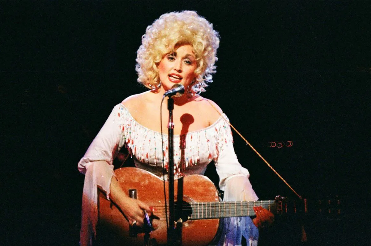 Dolly Parton sings into a microphone while she strums an acoustic guitar.