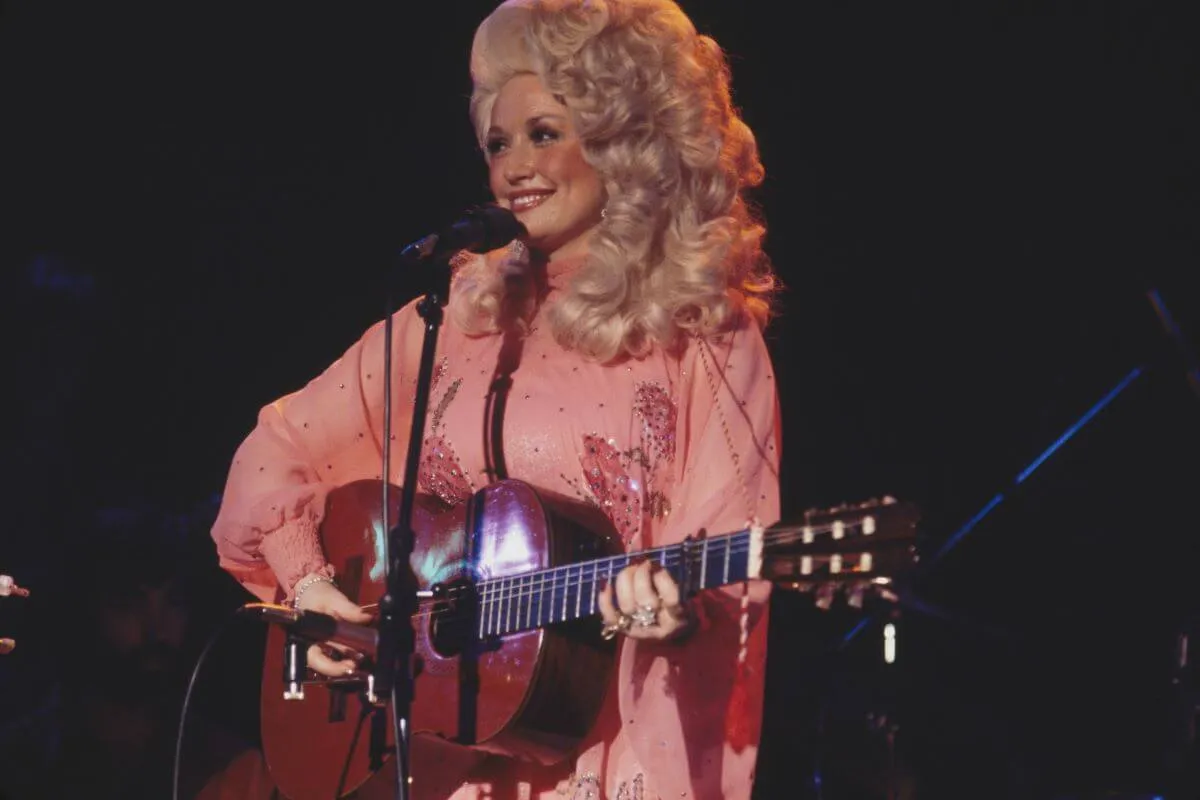 Dolly Parton wears a pink dress and plays guitar in front of a microphone.