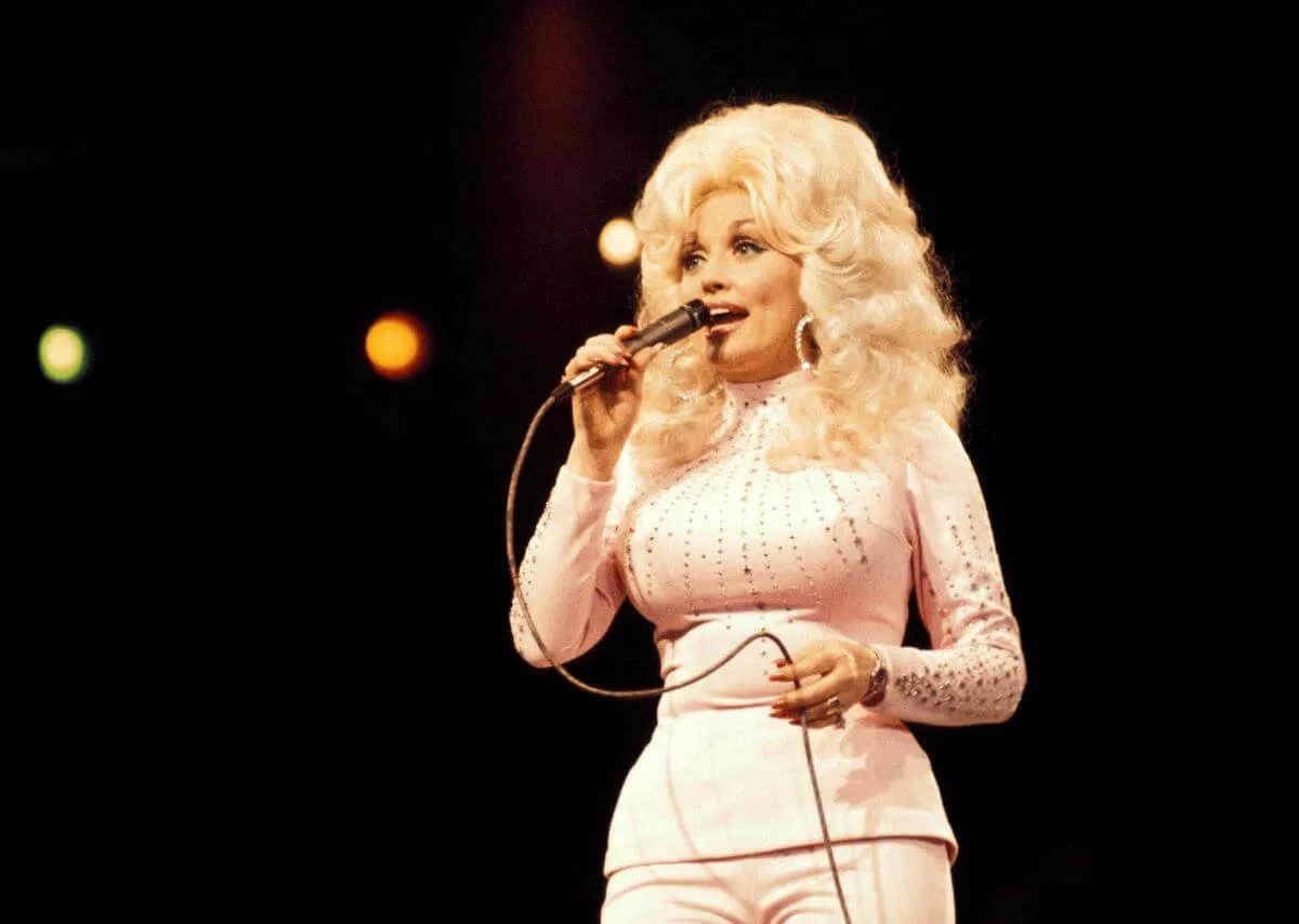 Dolly Parton wears a white shirt and white pants. She talks into a microphone.