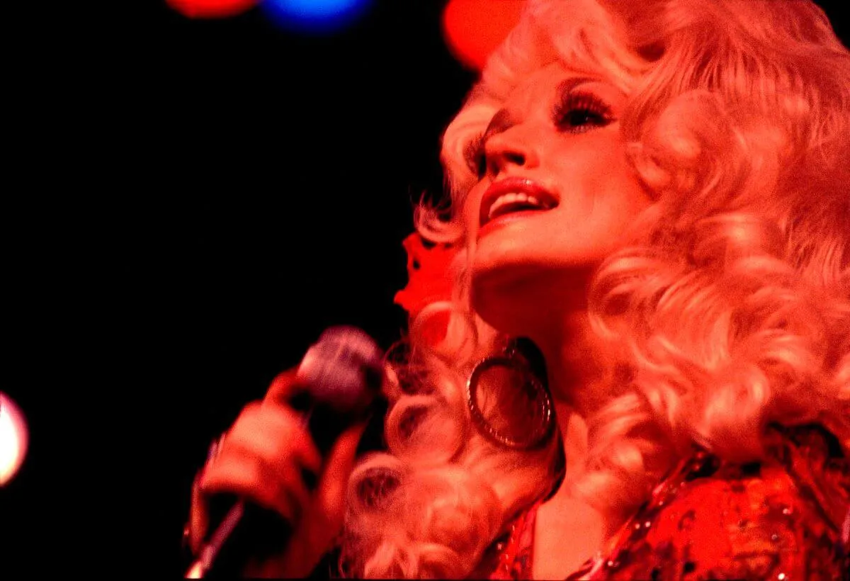 Dolly Parton sings into a microphone. She is lit with red light.