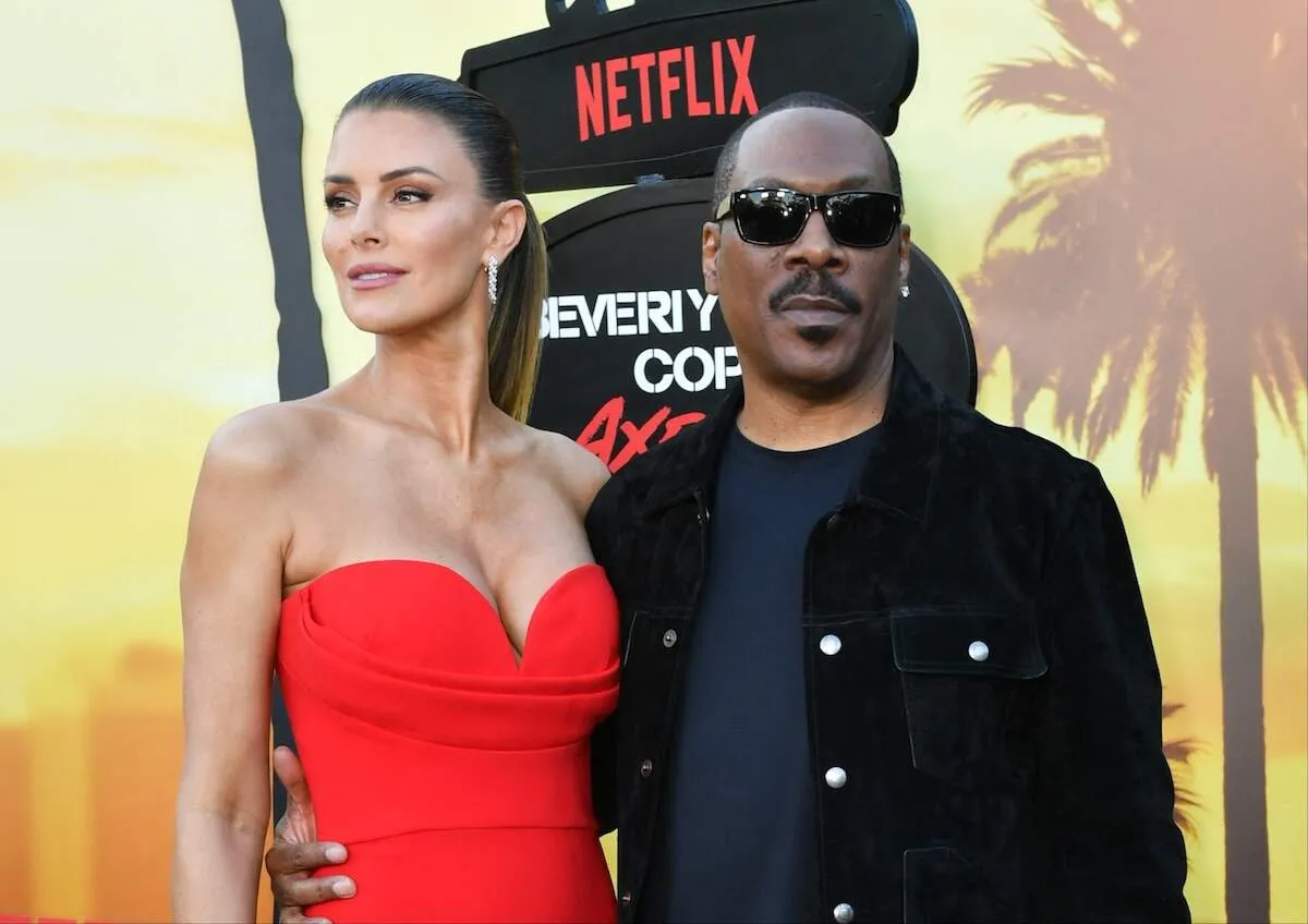 Wearing a black jacket, Eddie Murphy stands with his partner, Paige Butcher wearing a red dress, at the Beverly Hills Cop premiere