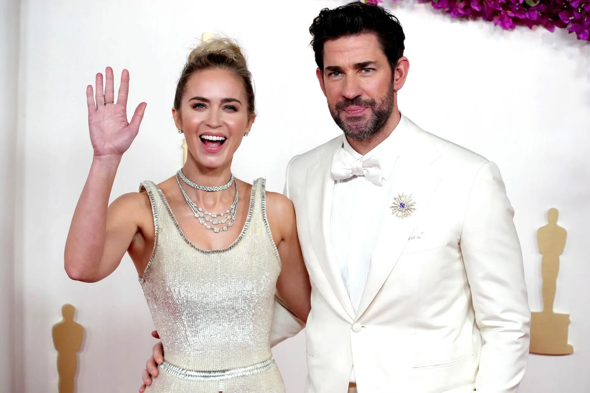 Emily Blunt waving while wearing a white dress and John Krasinski with his arm around her waist in a white suit at the 96th Annual Academy Awards