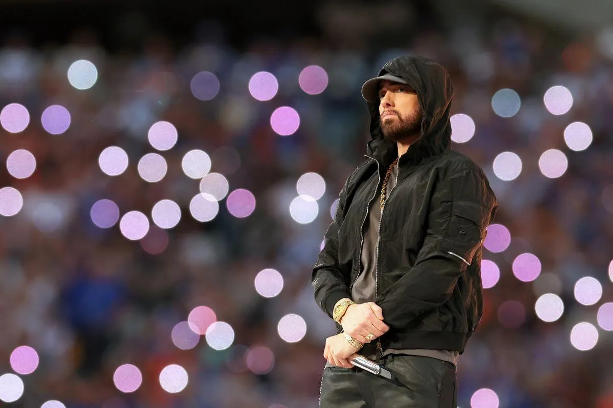 Eminem wears a black hoodie and hat. He stands on a stage and holds a microphone in front of him.