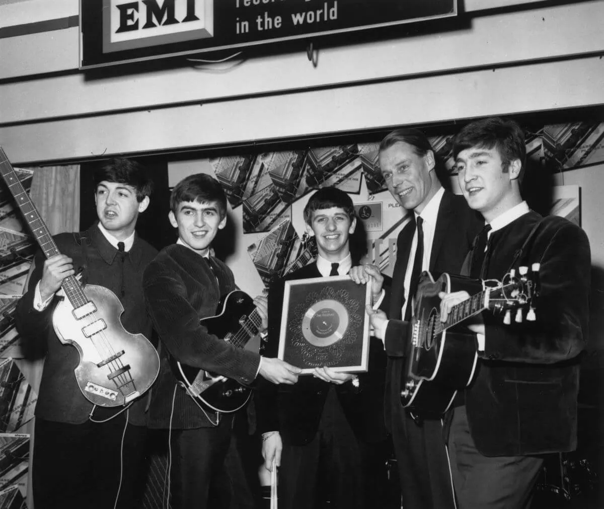 A black and white picture of The Beatles standing with George Martin. Paul McCartney, George Harrison, and John Lennon hold guitars. Ringo Starr holds a framed record.