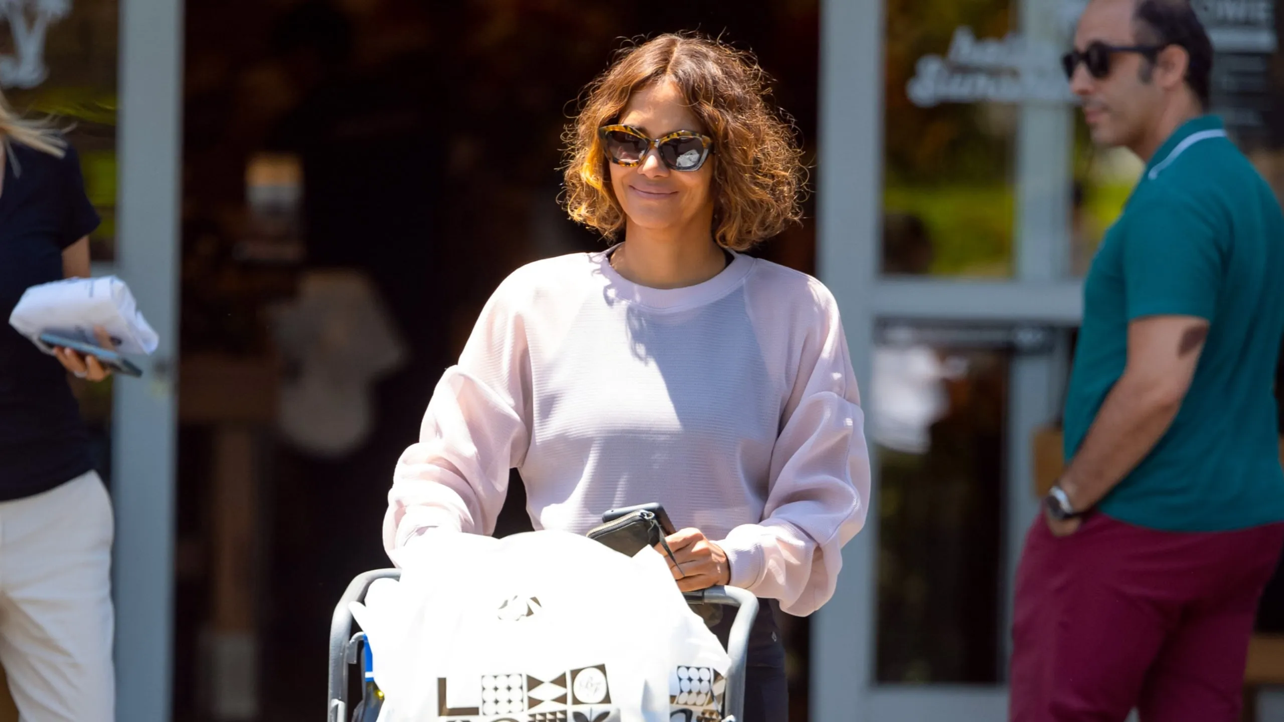 Wearing a pink sweatshirt and leggings, Hally Berry leaves a grocery store with a cart full of goods