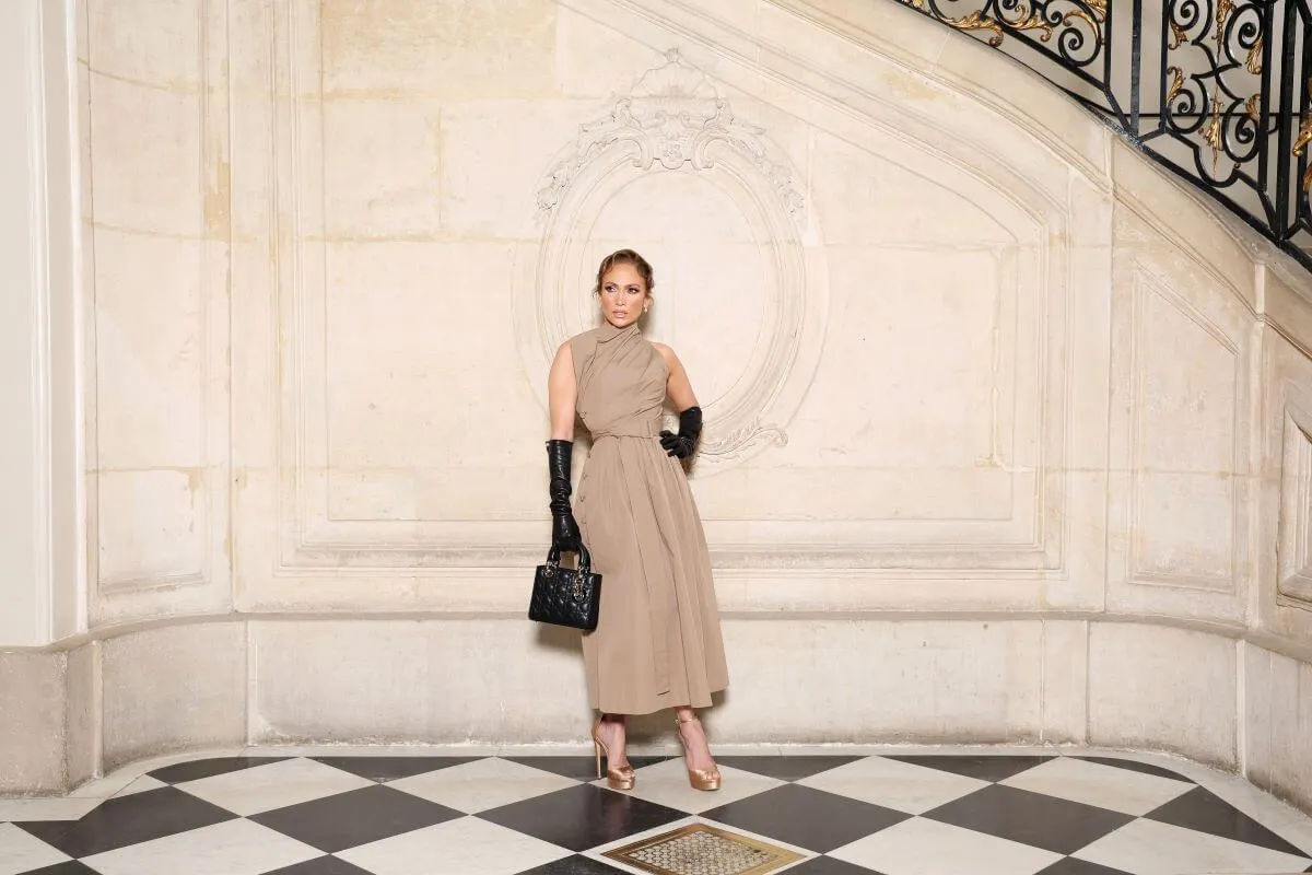 Jennifer Lopez wears a brown dress and black gloves. She stands in front of a staircase with her hand on her hip.