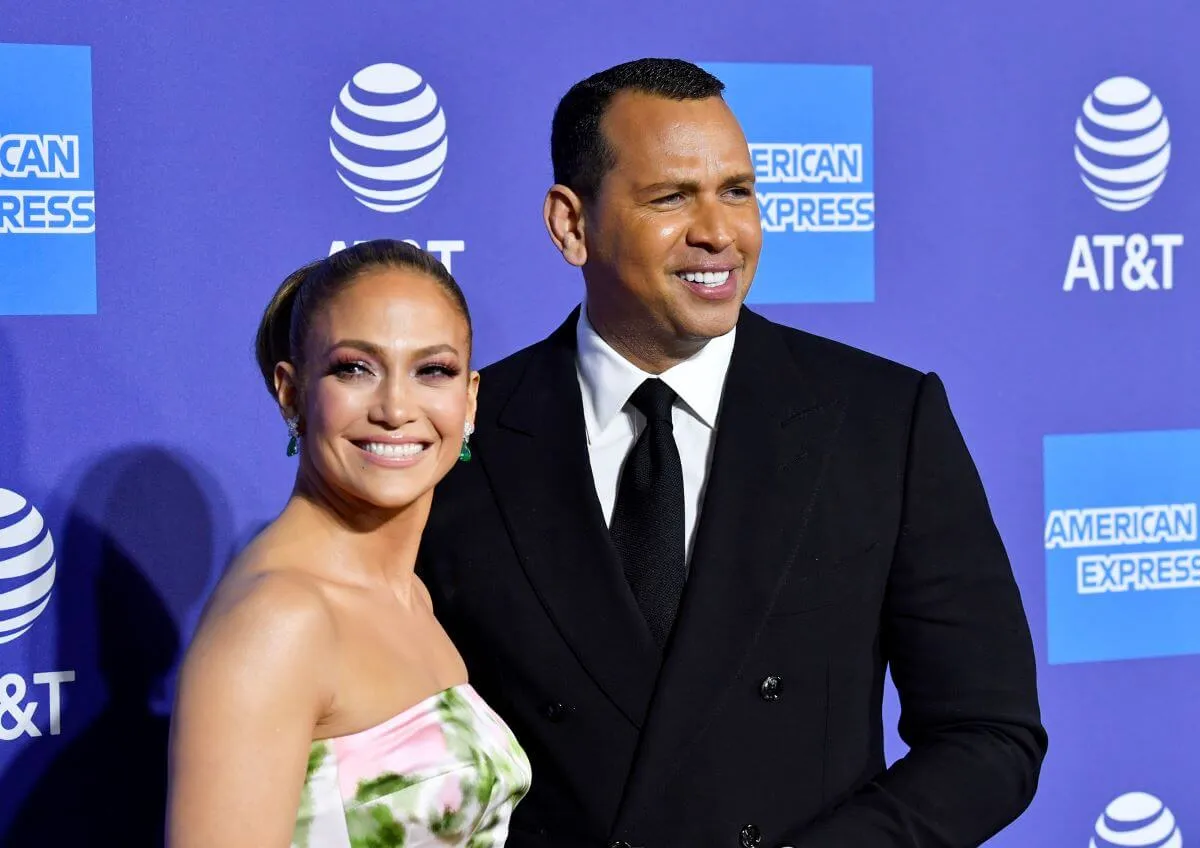 Jennifer Lopez wears a strapless dress and stands with Alex Rodriguez, who wears a suit. They are in front of a blue background.