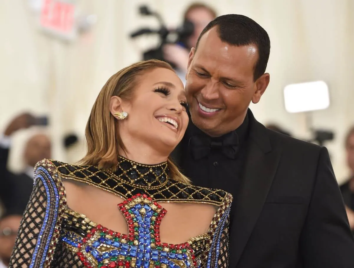 Jennifer Lopez wears a beaded dress with a colorful cross on her chest. She leans against Alex Rodriguez, who wears a black suit.