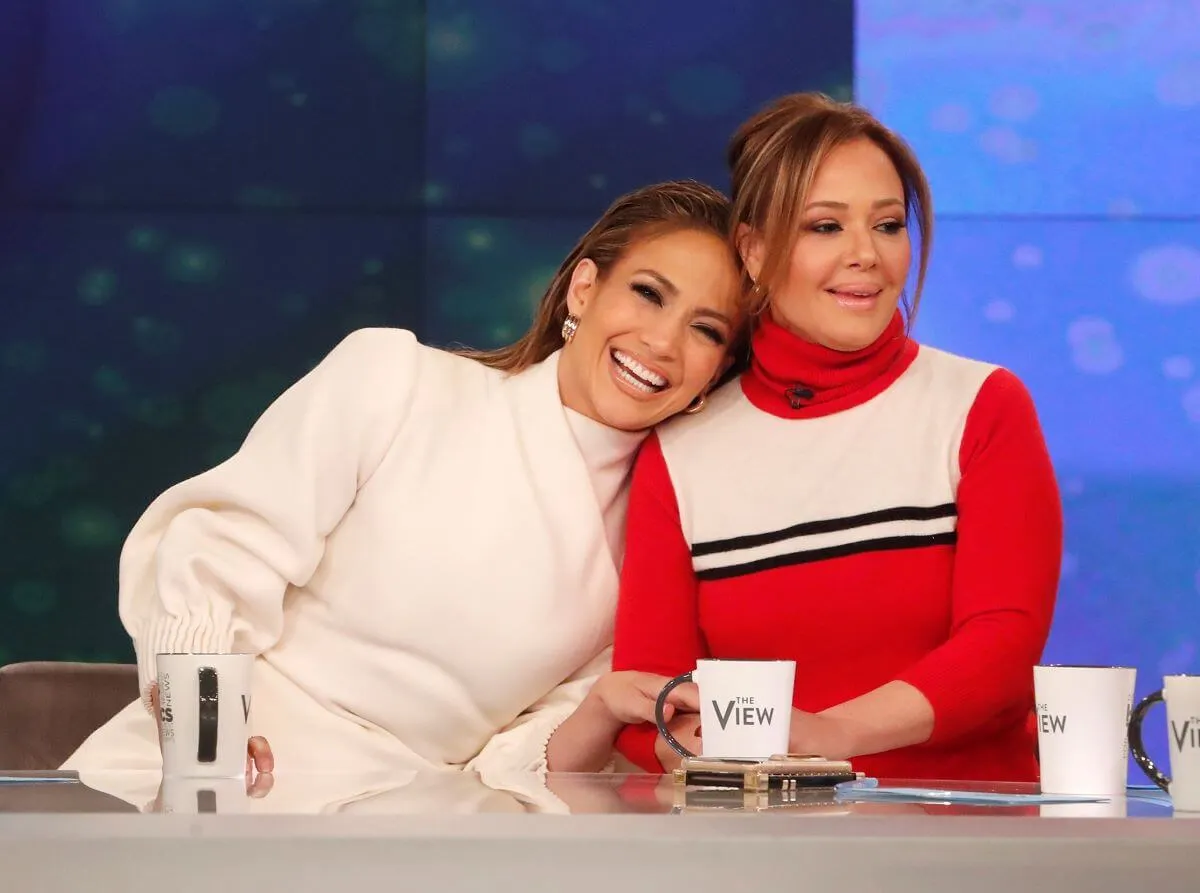 Jennifer Lopez leans on Leah Remini's shoulder. Lopez wears a white sweater and Remini wears a red and white sweater.