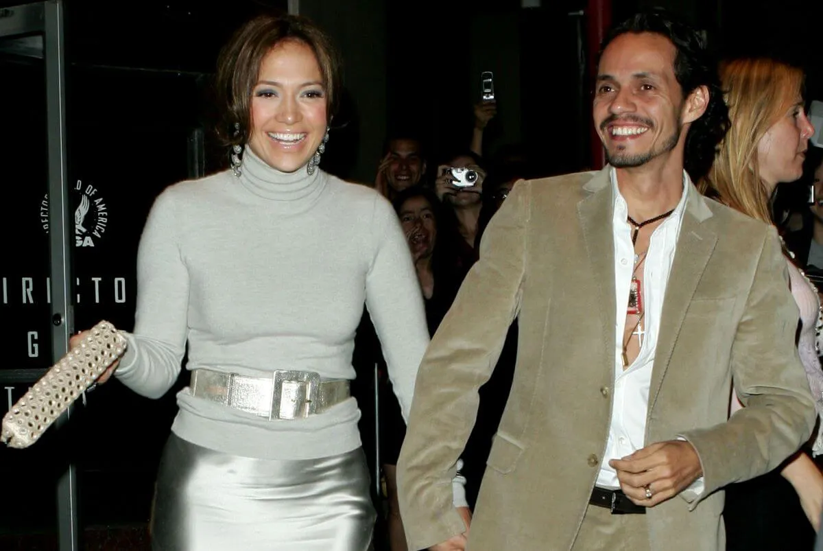 Jennifer Lopez and Marc Anthony hold hands as they walk outside. She wears a silver turtleneck with a matching belt and skirt. He wears a tan suit.