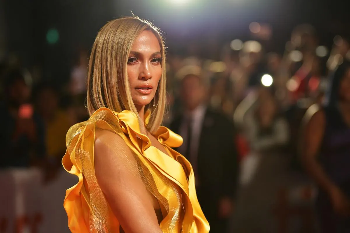 Jennifer Lopez wears a yellow dress and stands on a red carpet.