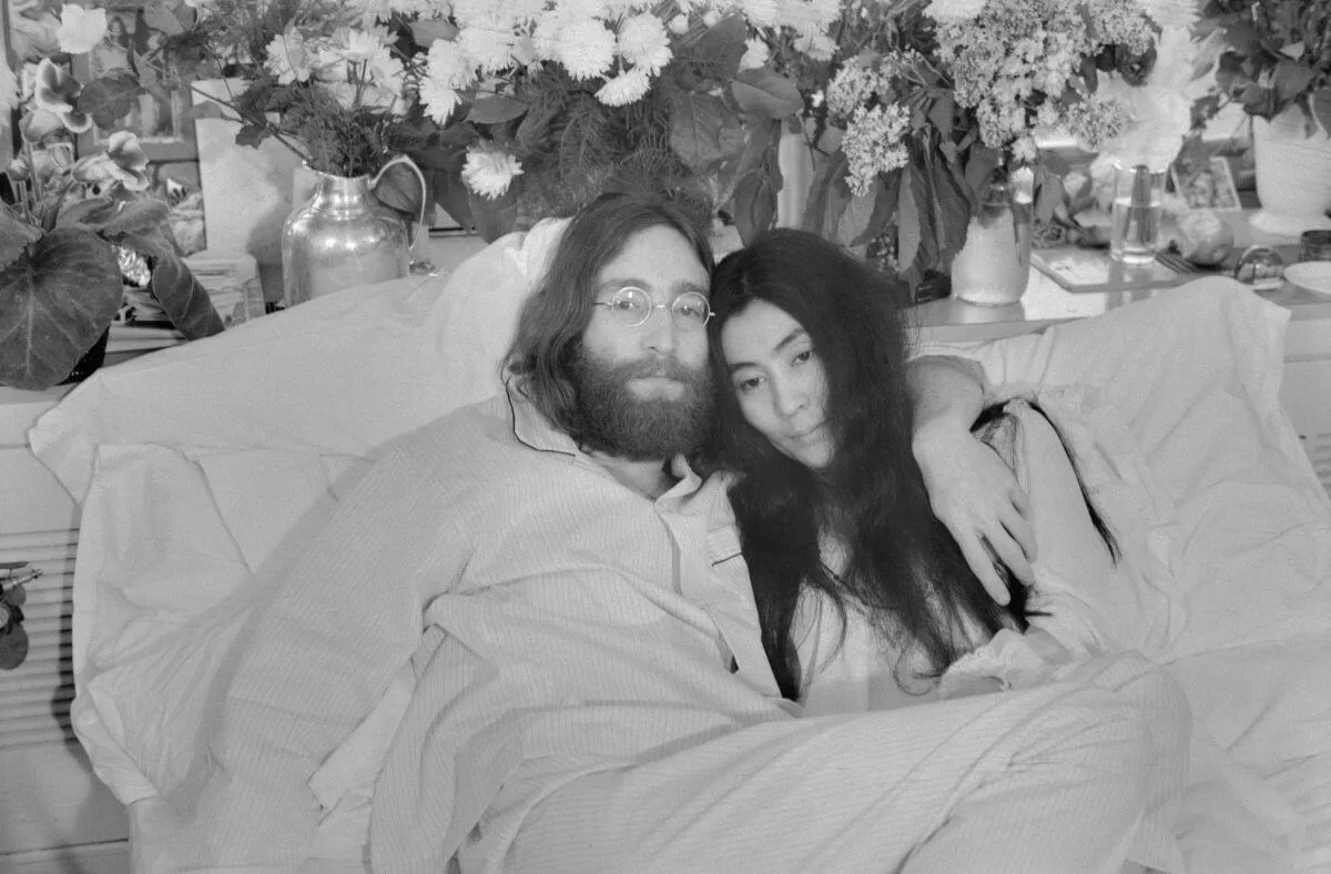 A black and white picture of John Lennon and Yoko Ono laying together in bed. He has his arm around her shoulders.