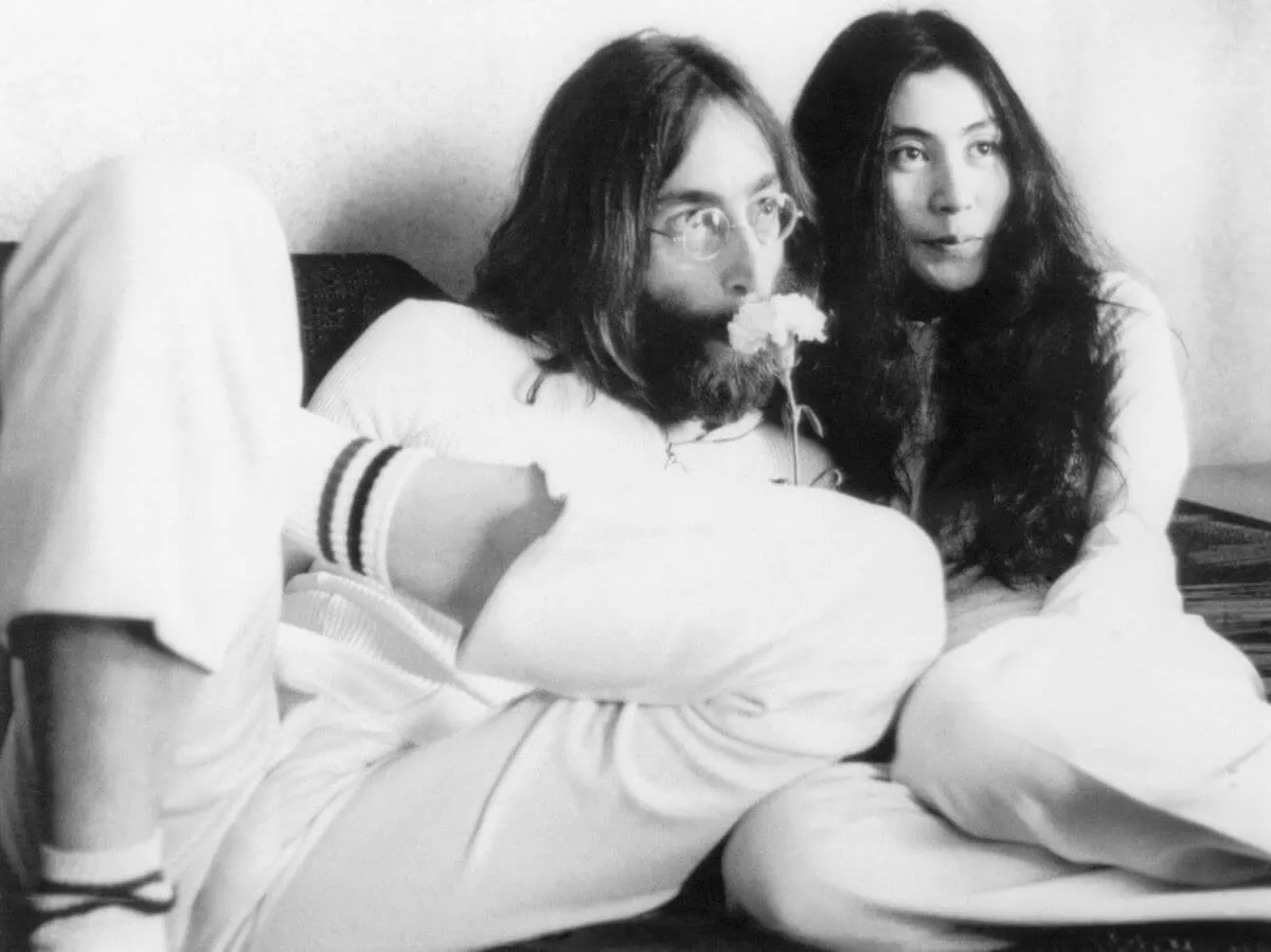 A black and white picture of John Lennon and Yoko Ono sitting in a bed together. Lennon sniffs a flower.