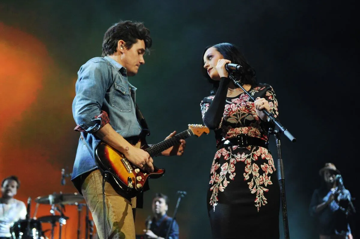 Katy Perry holds a microphone and looks at John Mayer, who plays guitar.