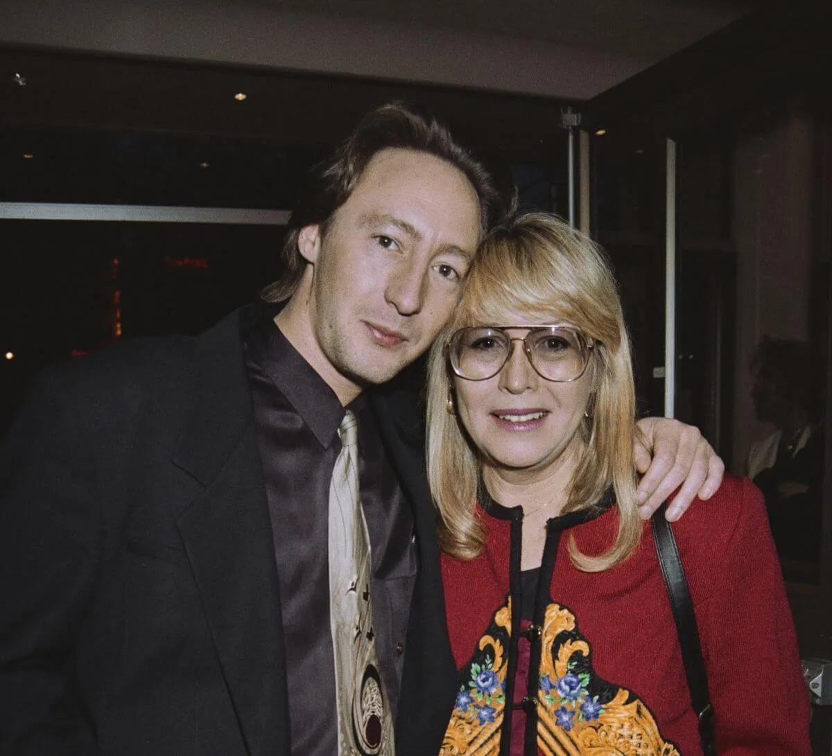 Julian Lennon stands with his arm around Cynthia Lennon's shoulders. She wears tinted glasses and a red sweater. He wears a shirt and tie. 