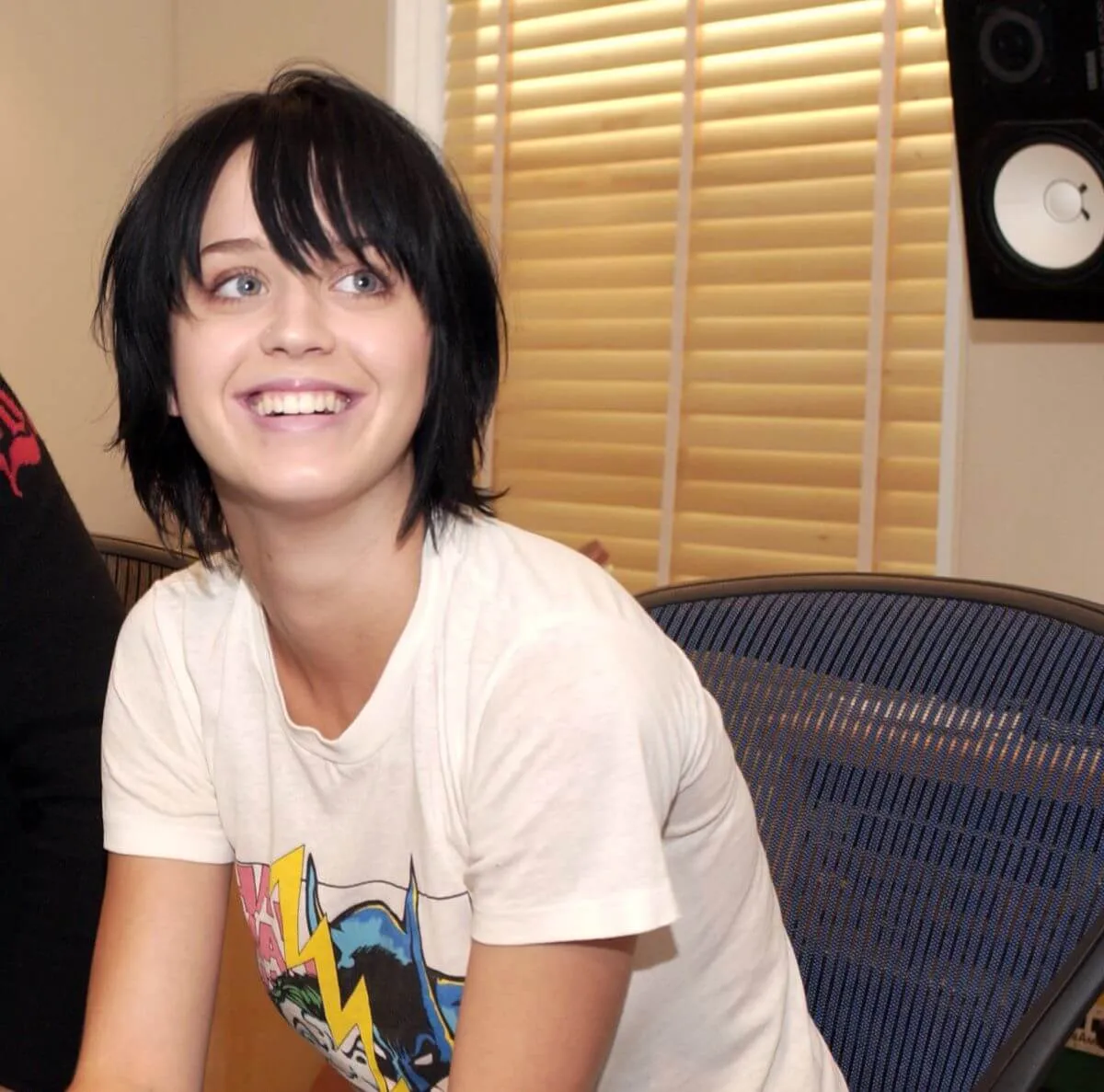 Katy Perry wears a white graphic shirt and sits on a couch in front of a window.