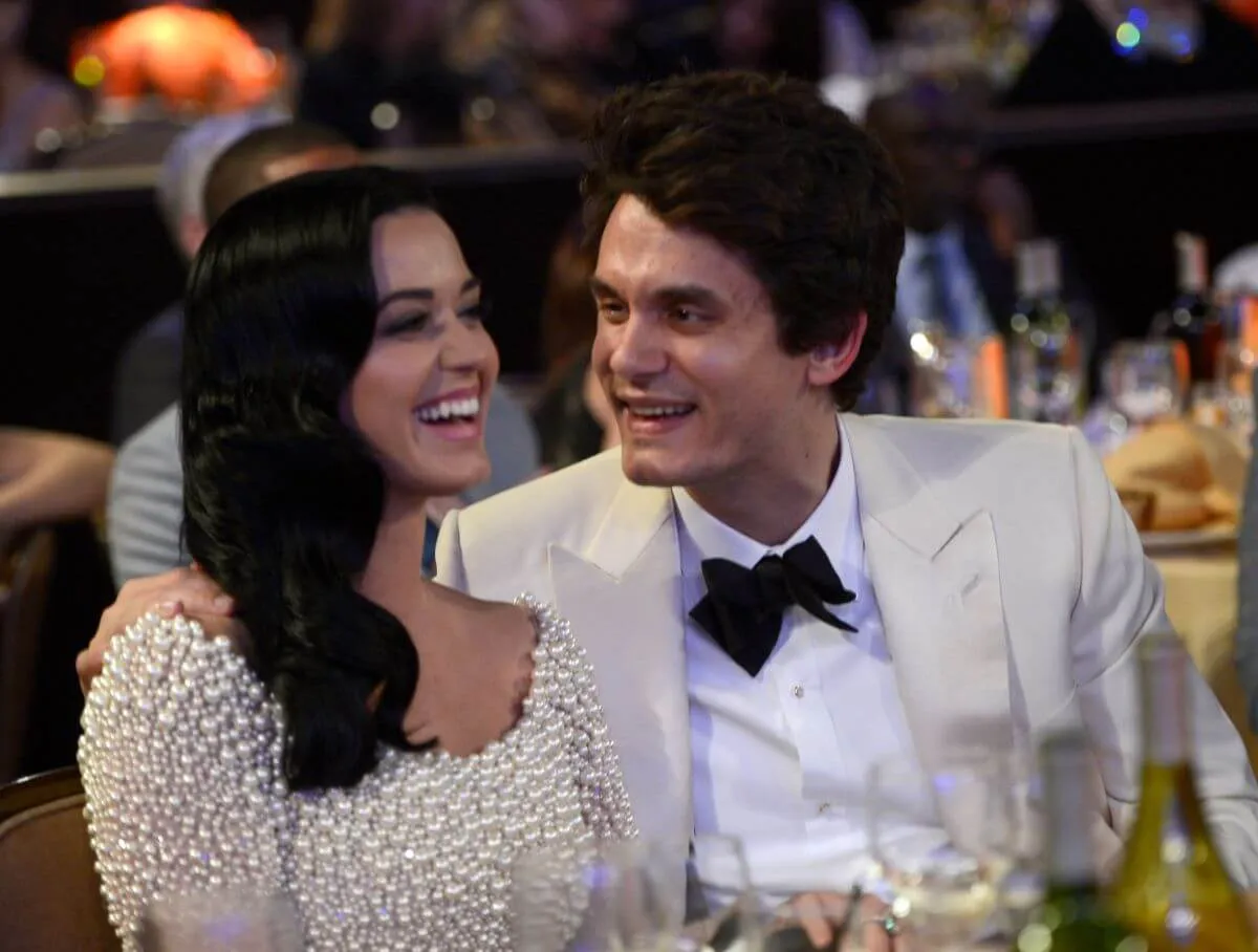 Katy Perry and John Mayer sit at a table together. He looks at her and they both laugh. She wears a white dress and he wears a white suit.