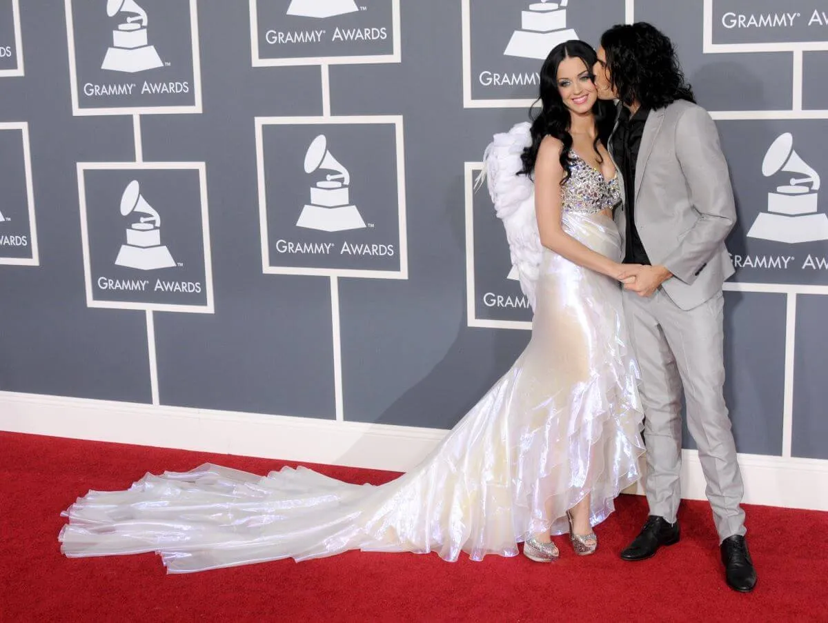 Katy Perry and Russell Brand stand together on the red carpet at the Grammys. He kisses her on the cheek and they hold hands. She wears a white dress and he wears a gray suit.