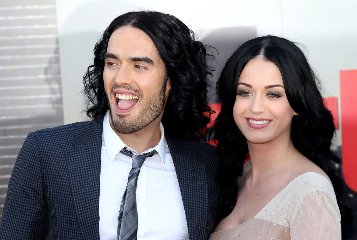 Russell Brand and Katy Perry stand together and smile. He wears a suit and she wears a white dress.
