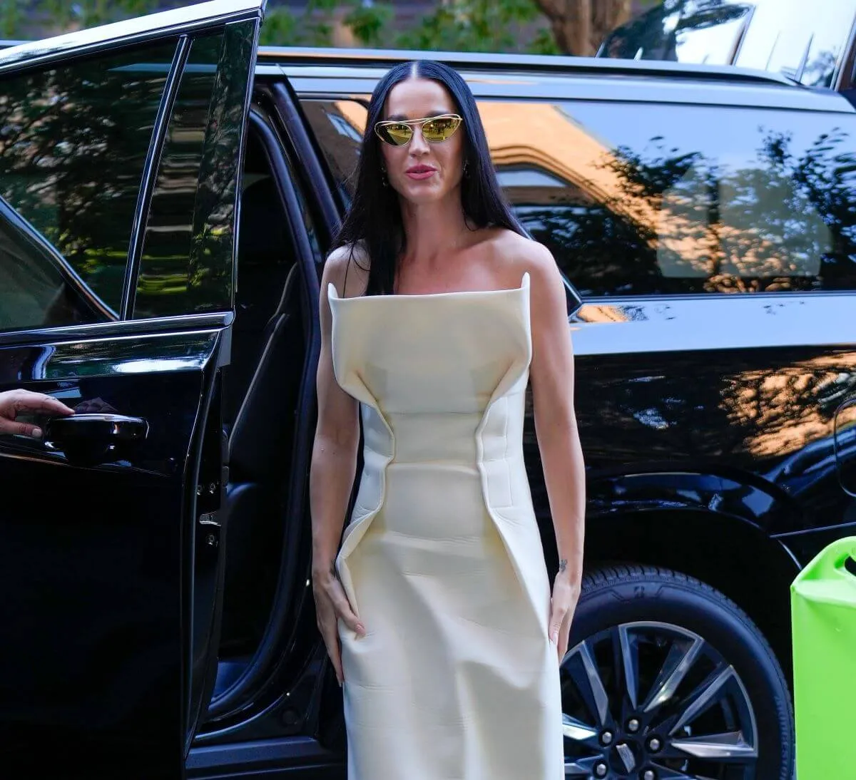 Katy Perry wears a white strapless dress and sunglasses. She stands next to the open door of a black SUV.