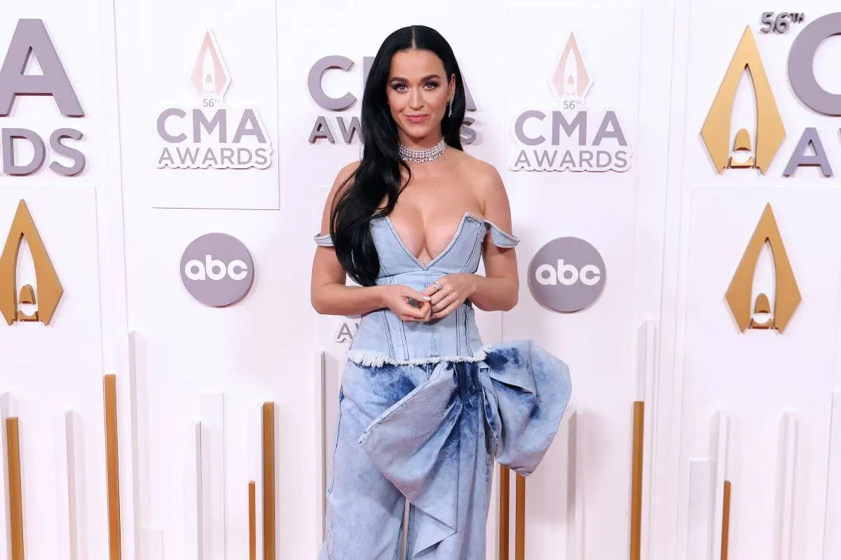 Katy Perry wears a light blue denim dress and stands in front of a step and repeat banner for the CMAs.