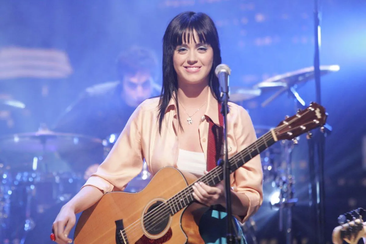 Katy Perry holds an acoustic guitar and stands in front of a microphone.