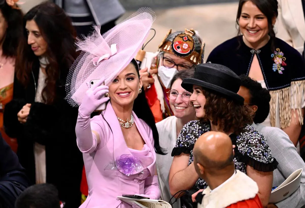 Katy Perry wears a lavender dress and matching hat. She takes a selfie with fans at  the Coronation of King Charles III and Queen Camilla.