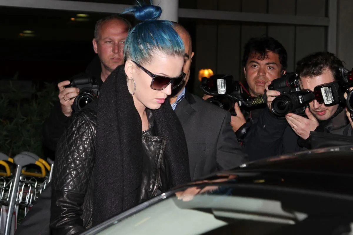 Katy Perry wears sunglasses and a black coat and walks through a crowd of paparazzi. She wears her blue hair in a bun.