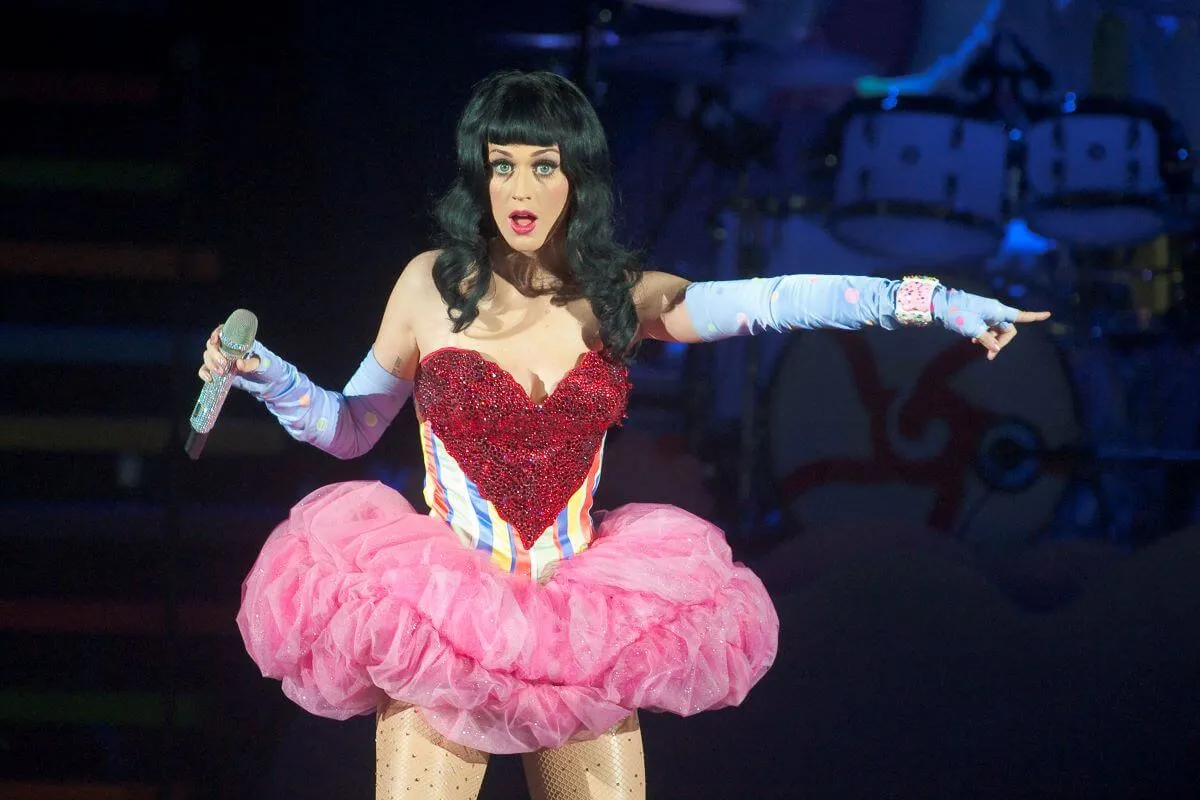 Katy Perry wears a pink tutu and stands on a stage. She holds a microphone in one hand and points her other hand.