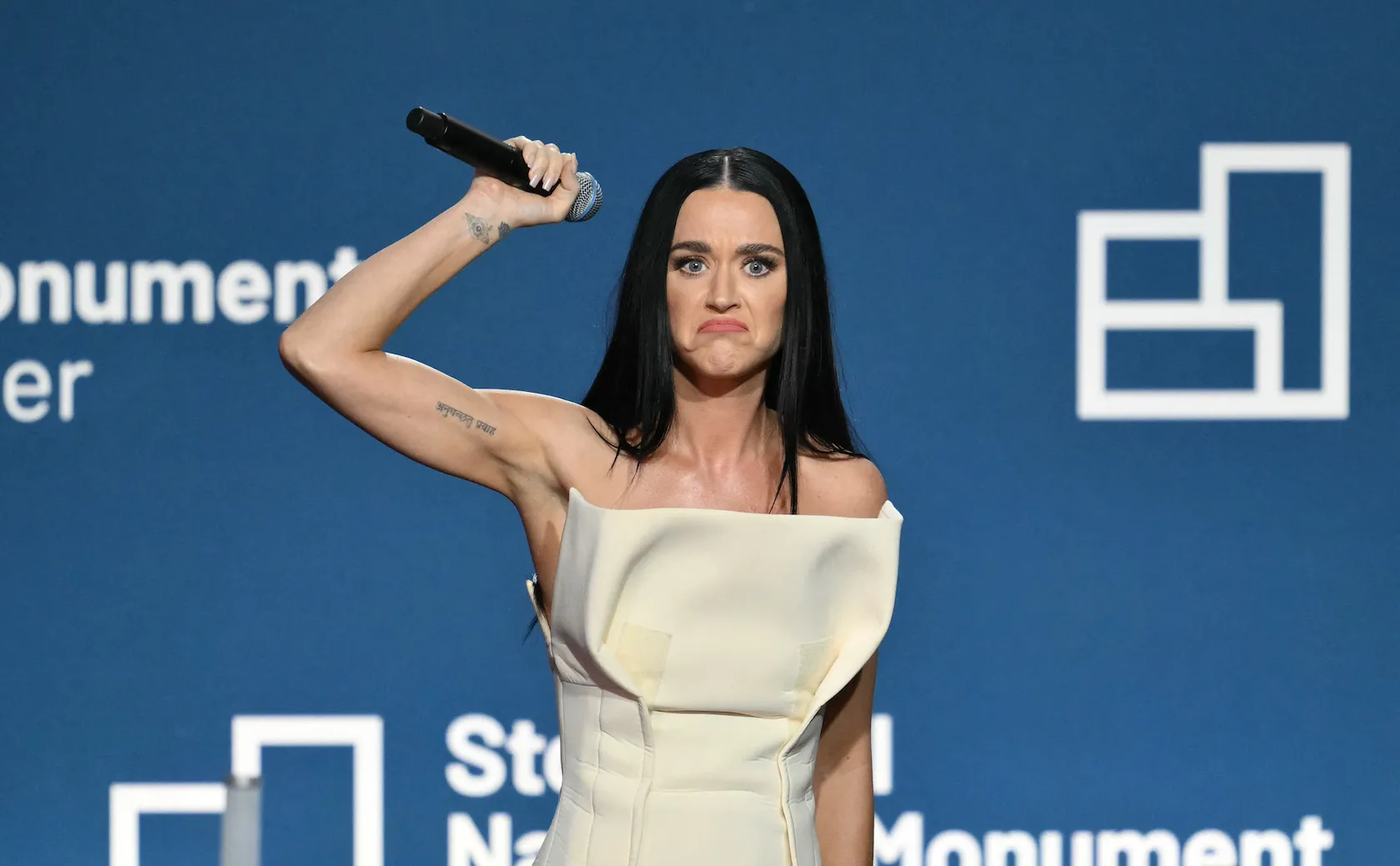 Katy Perry holding her arm up with a microphone while wearing a white strapless dress at an event