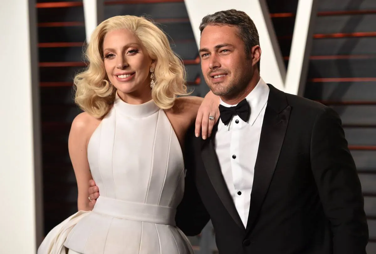 Lady Gaga wears a white dress and stands with her arm on Taylor Kinney's shoulder. He wears a tuxedo.