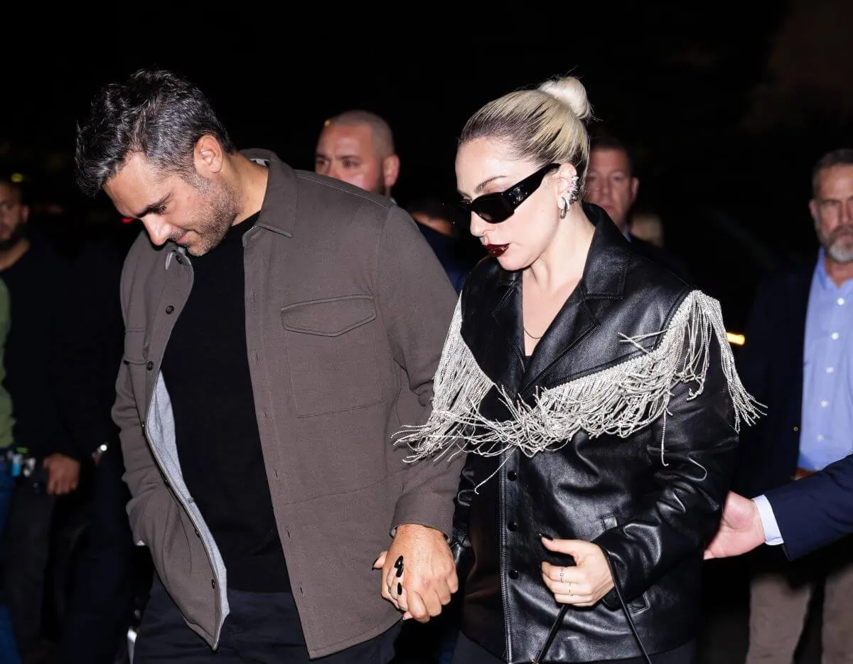 Lady Gaga and her fiancé Michael Polansky hold hands and walk together. He wears a gray jacket and she wears a black leather jacket and sunglasses.