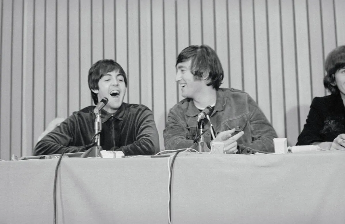 A black and white picture of Paul McCartney and John Lennon sitting behind a table and laughing. They have microphones in front of them.