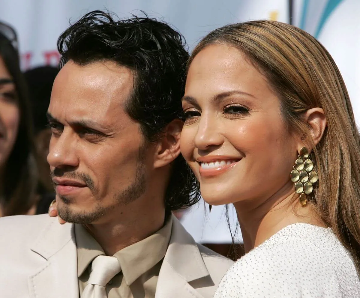 Jennifer Lopez smiles at the camera while standing with her arm around Marc Anthony's shoulder. She wears white and he wears a cream colored suit.