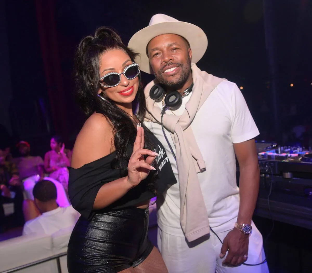 Mya wears sunglasses, a black shirt, and black shorts. She holds up a peace sign and stands with D-Nice, who wears a hat, white shirt, white pants, and beige sweater around his shoulders.