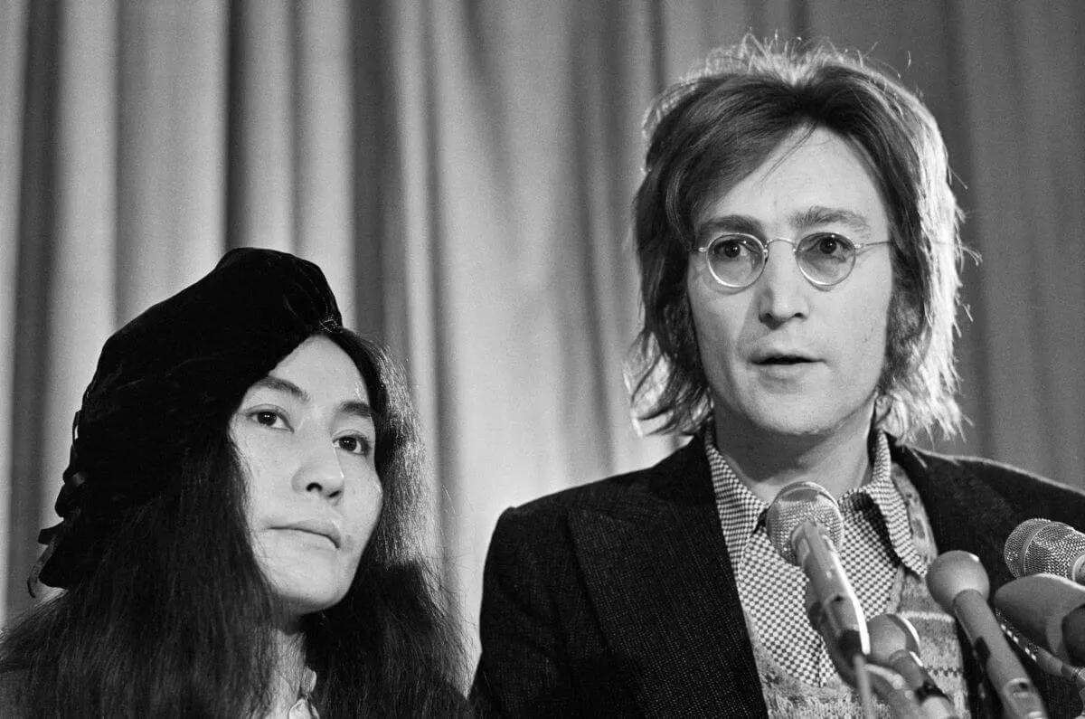 A black and white picture of Yoko Ono standing next to John Lennon. He stands in front of several microphones.