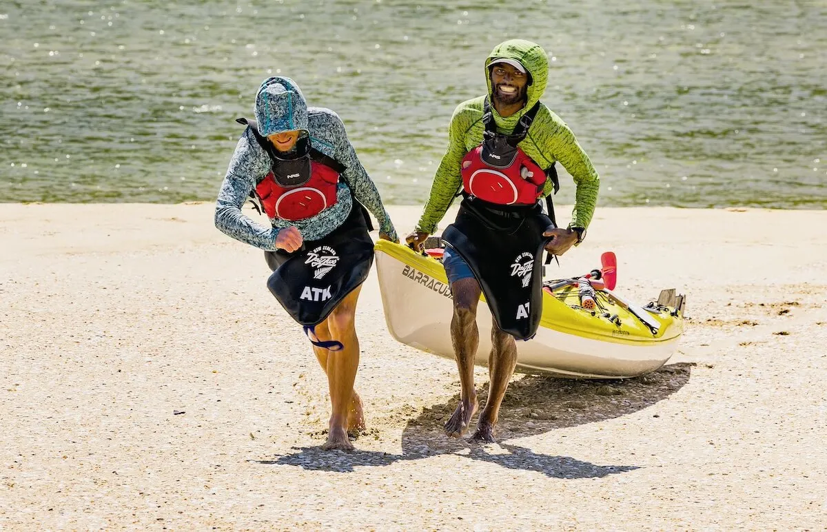 Two men pulling a kayak behind them on a beach in 'Race to Survive: New Zealand'